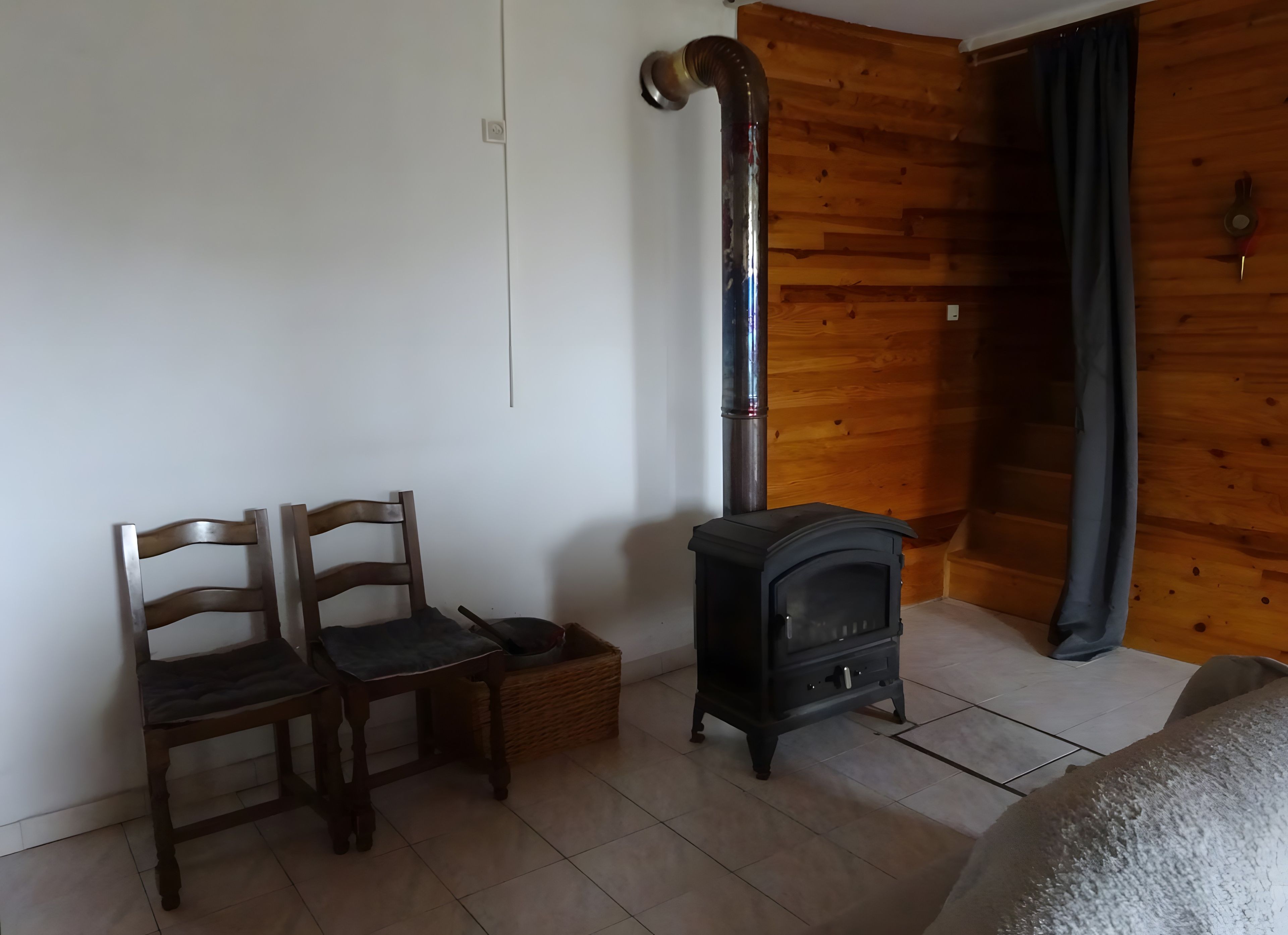 A room with a wooden wall, a black stove, two wooden chairs, and a small table.