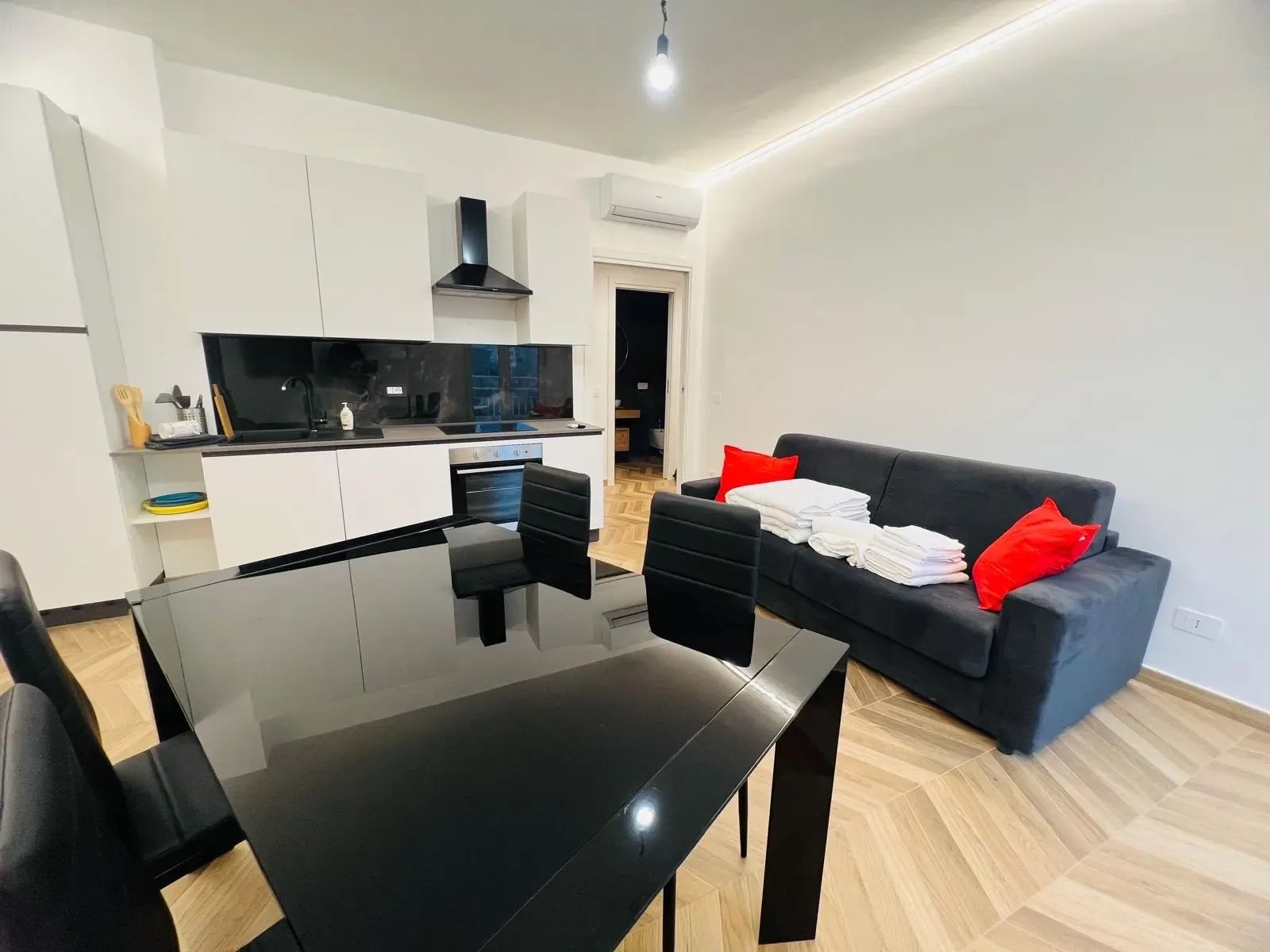 A modern living space with a dining area, kitchen, and a black sofa with red cushions.