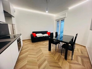 A modern kitchen with a built-in stove, a dining area with a glass table and chairs, a black sofa with red cushions, and a large window offering a city view. - New Listing (Sanremo)