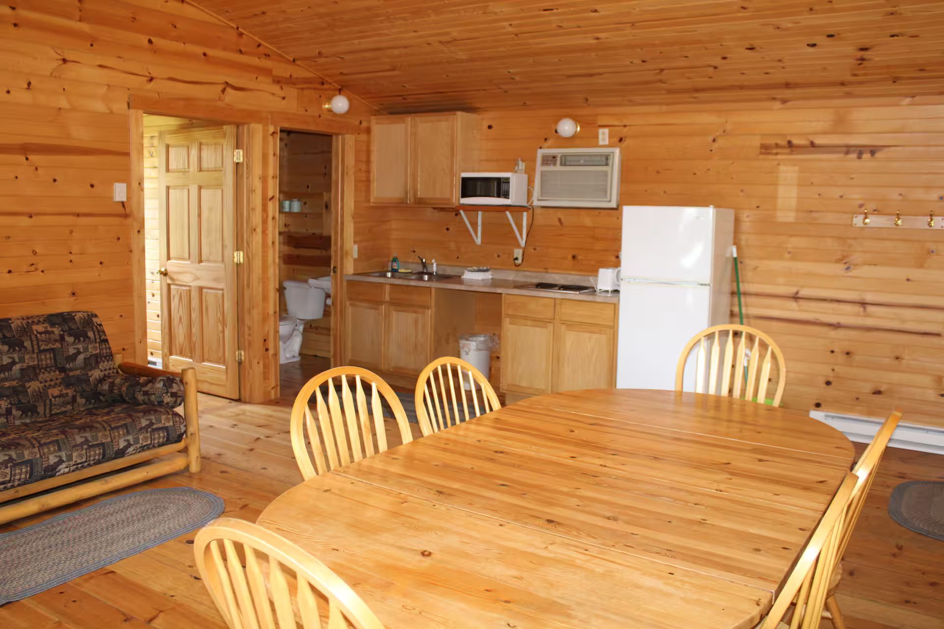 A wooden cabin interior with a dining table, chairs, a sofa, a kitchen area with appliances, and a door leading to another room.
