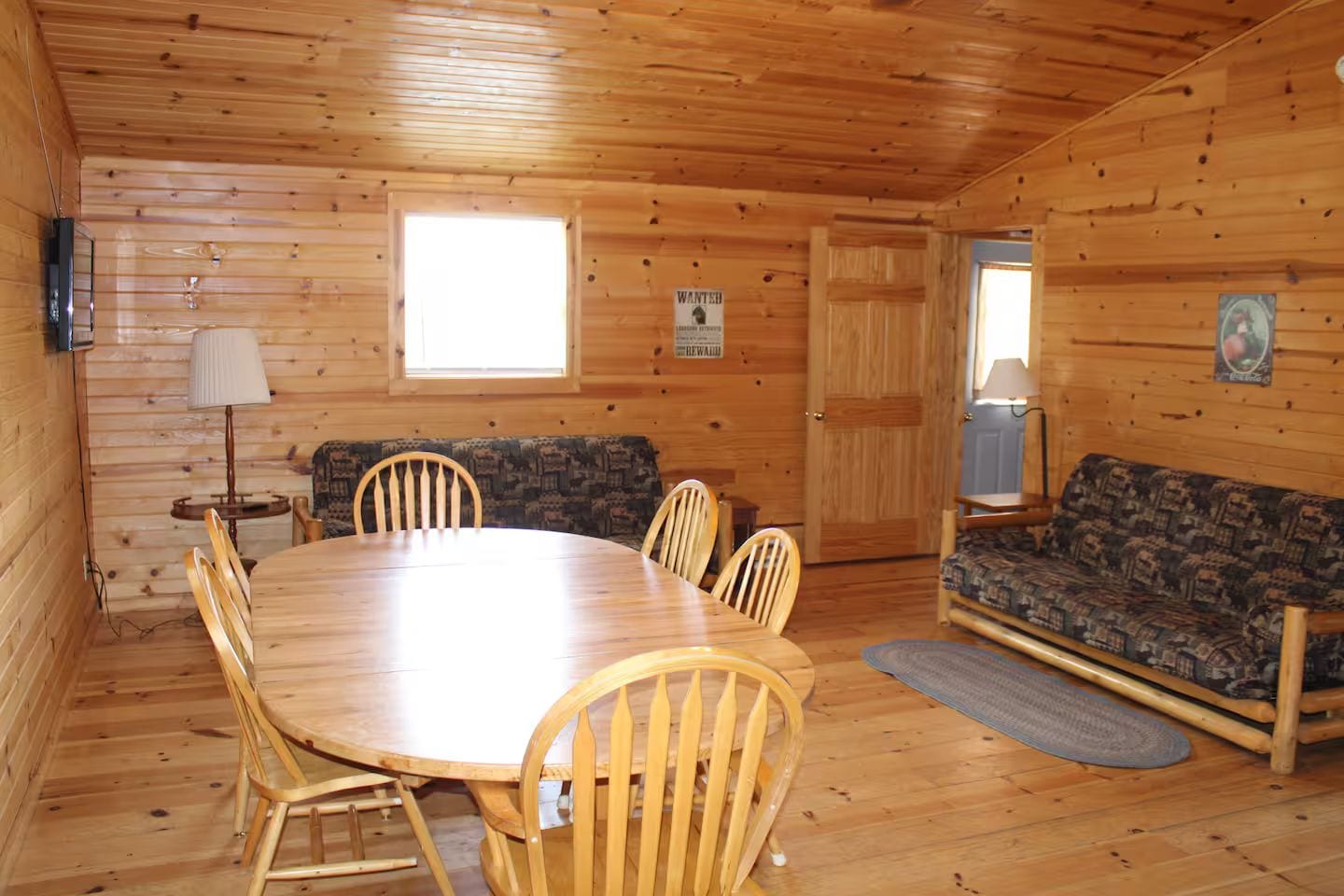 A wooden interior with a round table, four chairs, a sofa, a lamp, and a door.