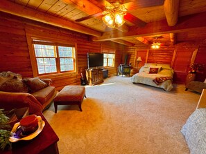 L’intérieur d’une cabane en rondins, avec un canapé, un pouf, une télévision, une cheminée et un lit.