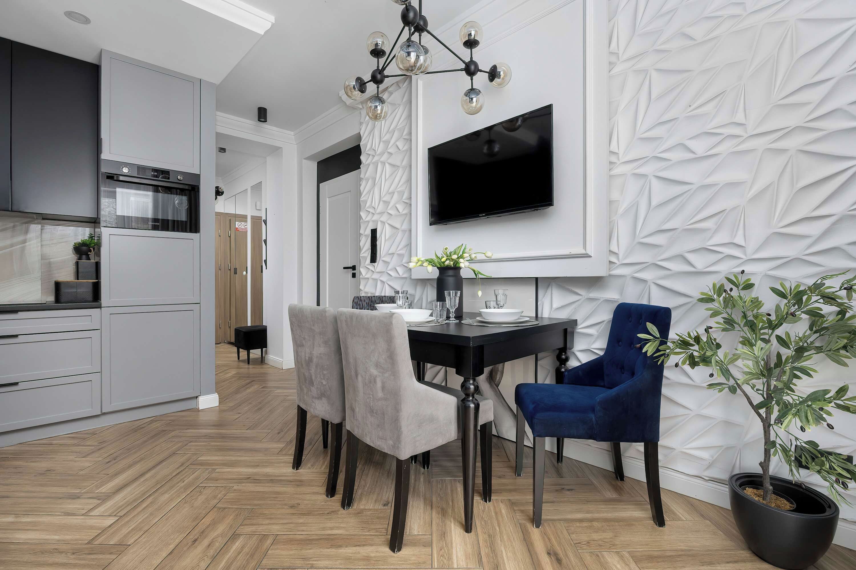 Eine moderne Küche mit eingebautem Ofen, eine Essecke mit einem schwarzen Tisch und grauen Stühlen sowie ein an der Wand befestigter Fernseher.
