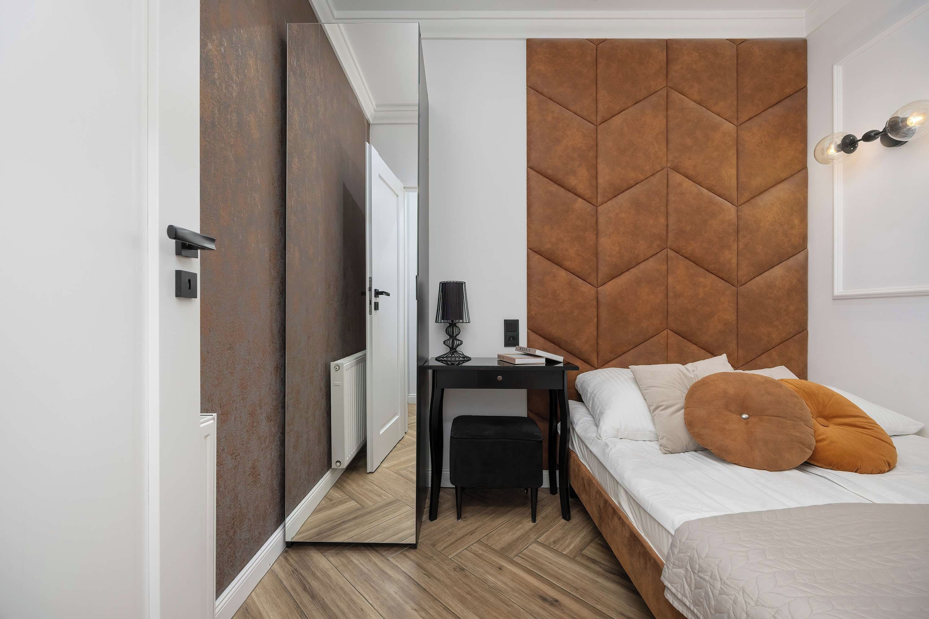Een moderne slaapkamer met een bruin hoofdeinde, een nachtkastje met een lamp, een zwarte stoel en een bed met witte en oranje kussens.