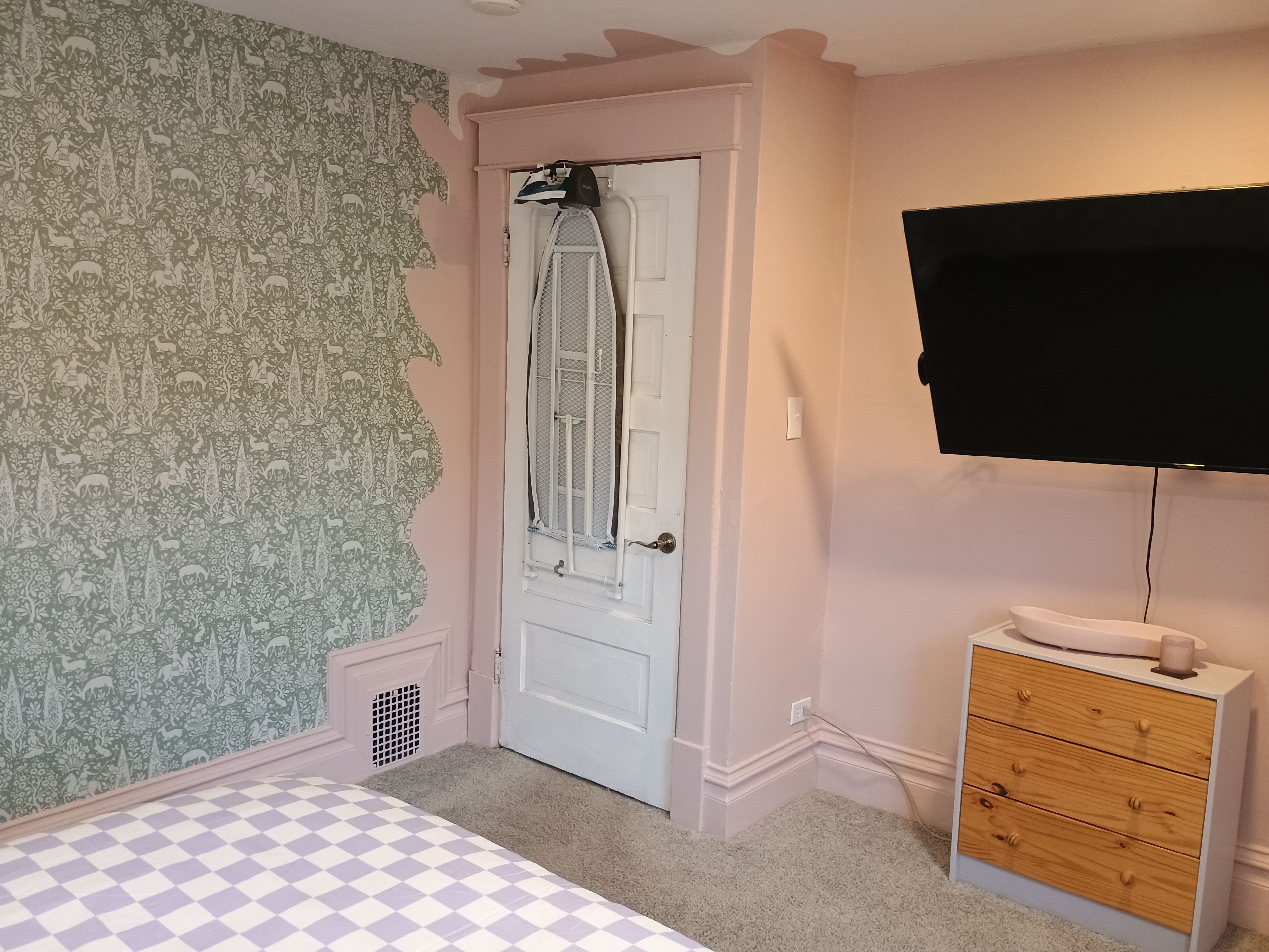 A bedroom with a checkered bedspread, a white door with a window, a television mounted on the wall, and a wooden dresser.