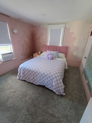 Une chambre à coucher avec une couvre-lit à carreaux, une tête de lit rose et une fenêtre avec des stores.