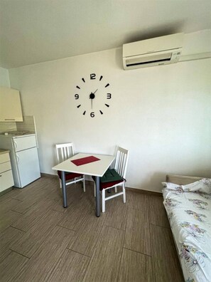 Ein kleiner, sauberer Raum mit einer weißen Wanduhr, einem Tisch mit zwei Stühlen, einem Bett mit Blumenmuster-Bettwäsche und einer Küchenzeile.
