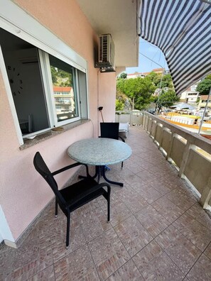 Ein Balkon mit rundem Tisch und zwei Stühlen, ein Fenster mit Jalousien und Blick auf die Stadt.
