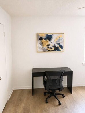 Um quarto com uma escrivaninha preta e cadeira de escritório, uma pintura abstrata emoldurada na parede e piso de madeira.