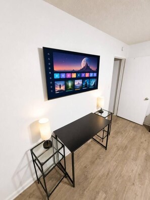 Quarto com televisão na parede exibindo um papel de parede com diversas imagens, uma mesa preta com duas luminárias e piso de madeira.