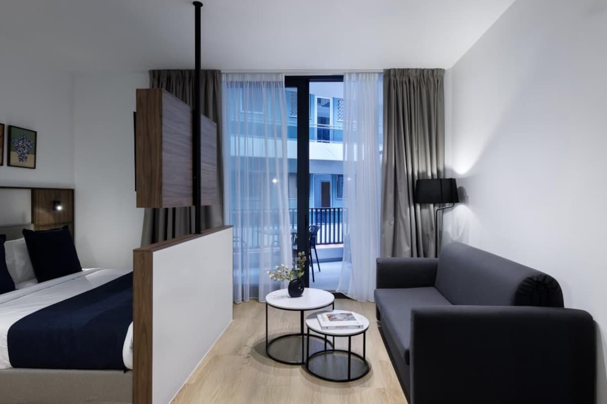 Habitación de hotel moderna con cama, sofá, una mesita con un florero y vistas a un balcón.
