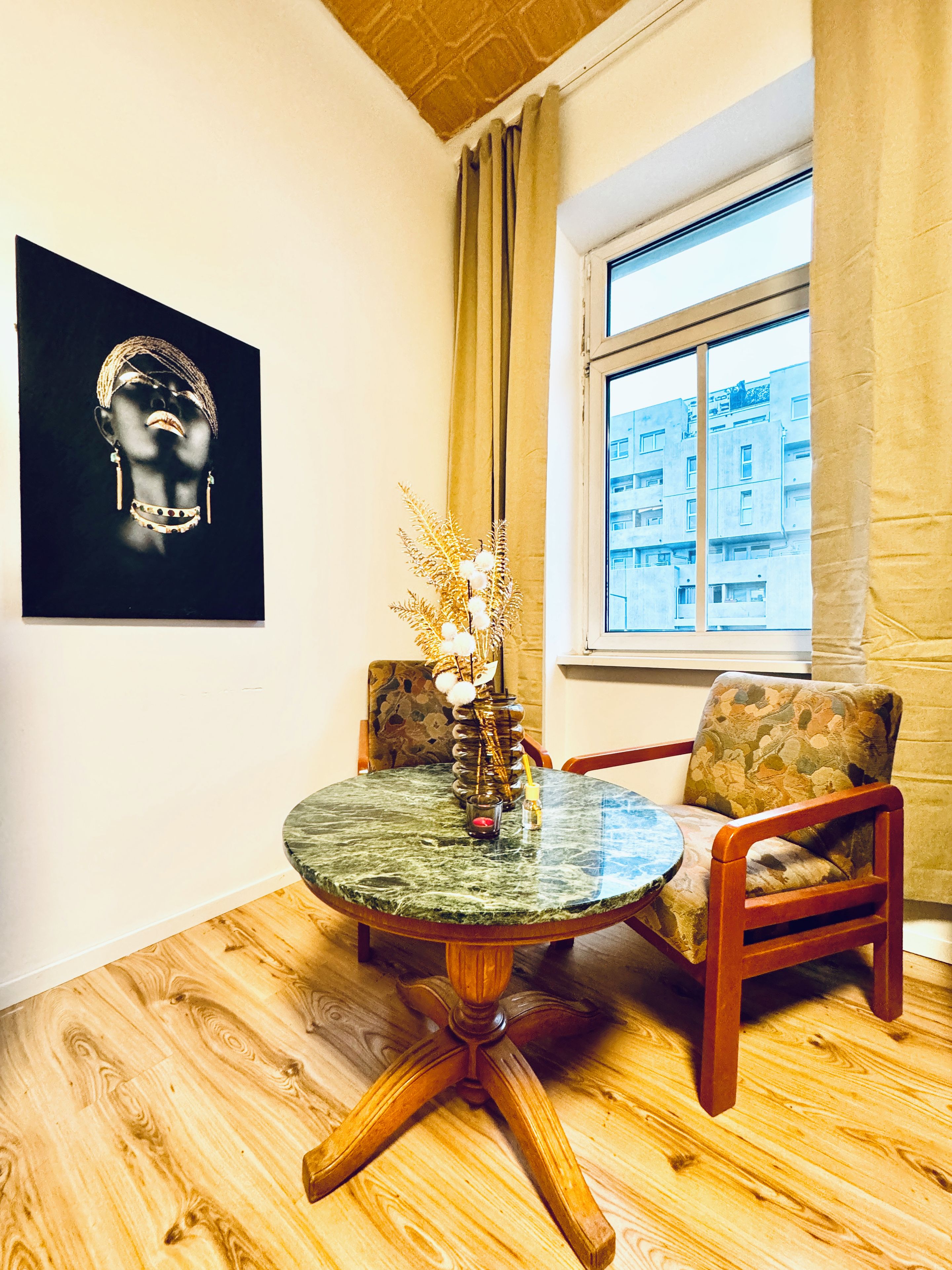 A room with a round table, two chairs, a window with curtains, a wall-mounted artwork, and a wooden floor.