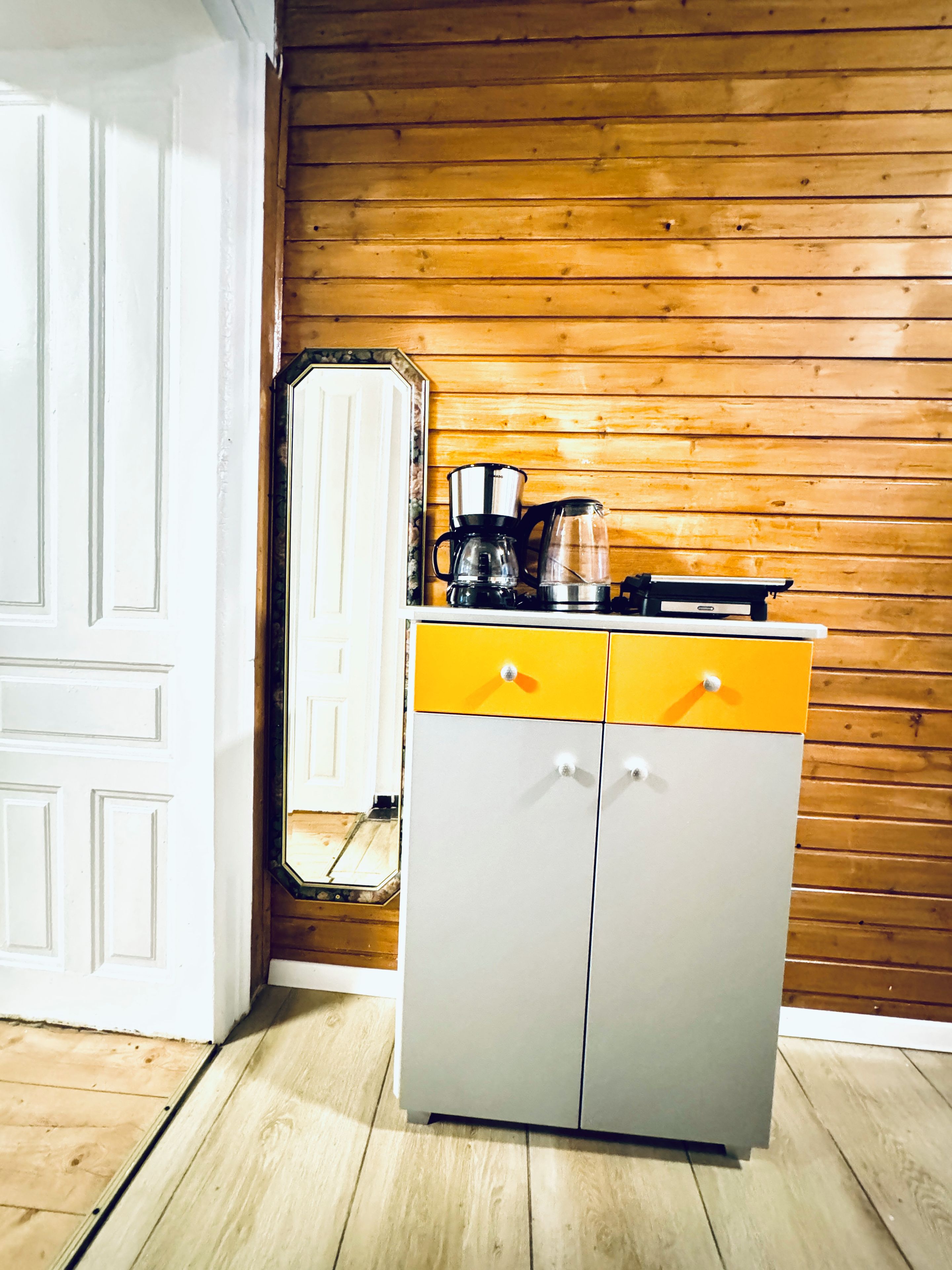 Een keuken met een houten wand, een witte deur en een geel-grijs kastje met een koffiezetapparaat erop.