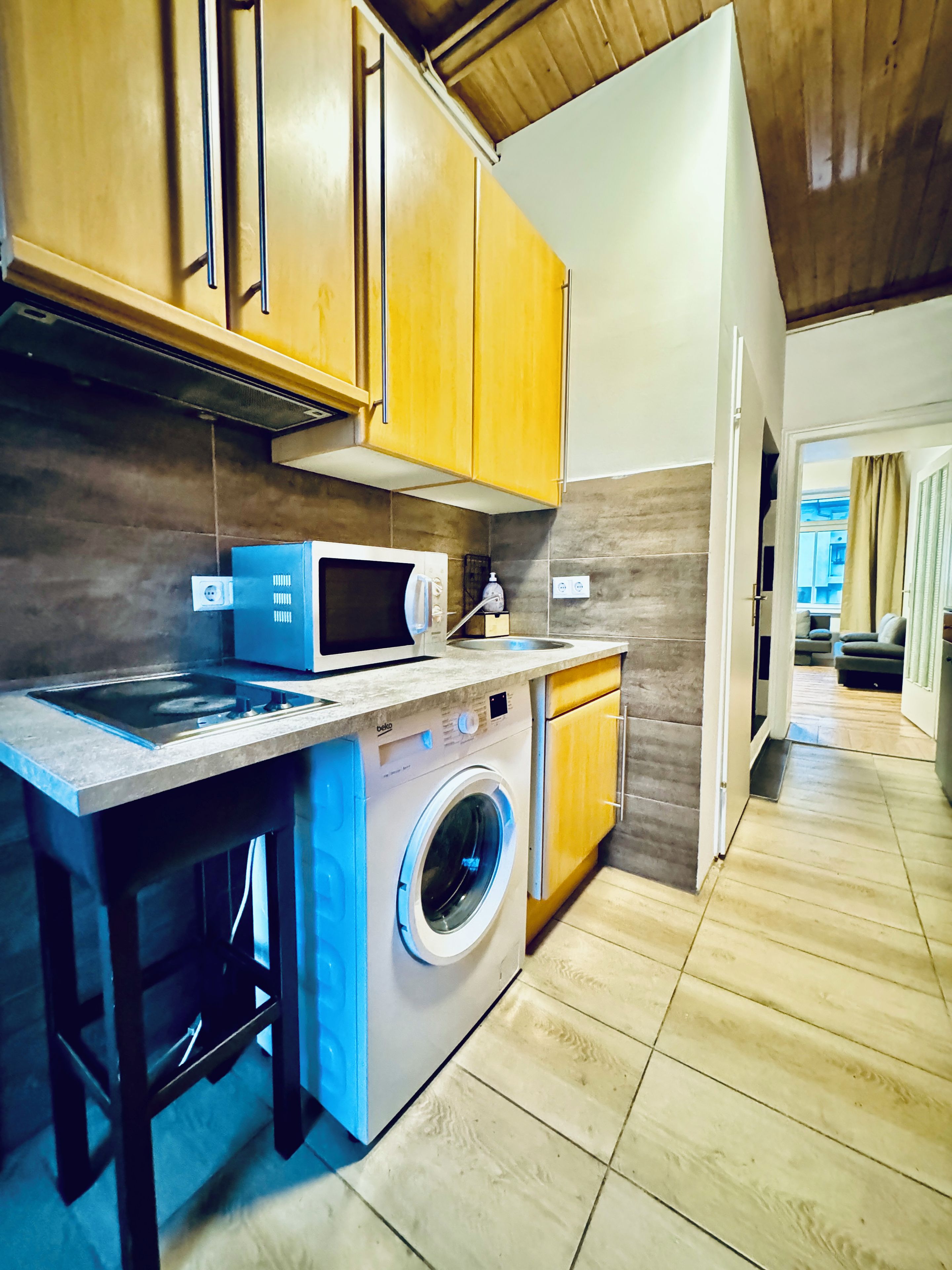 Een kleine keuken met houten kastjes, een magnetron, een wasmachine en een spoelbak.