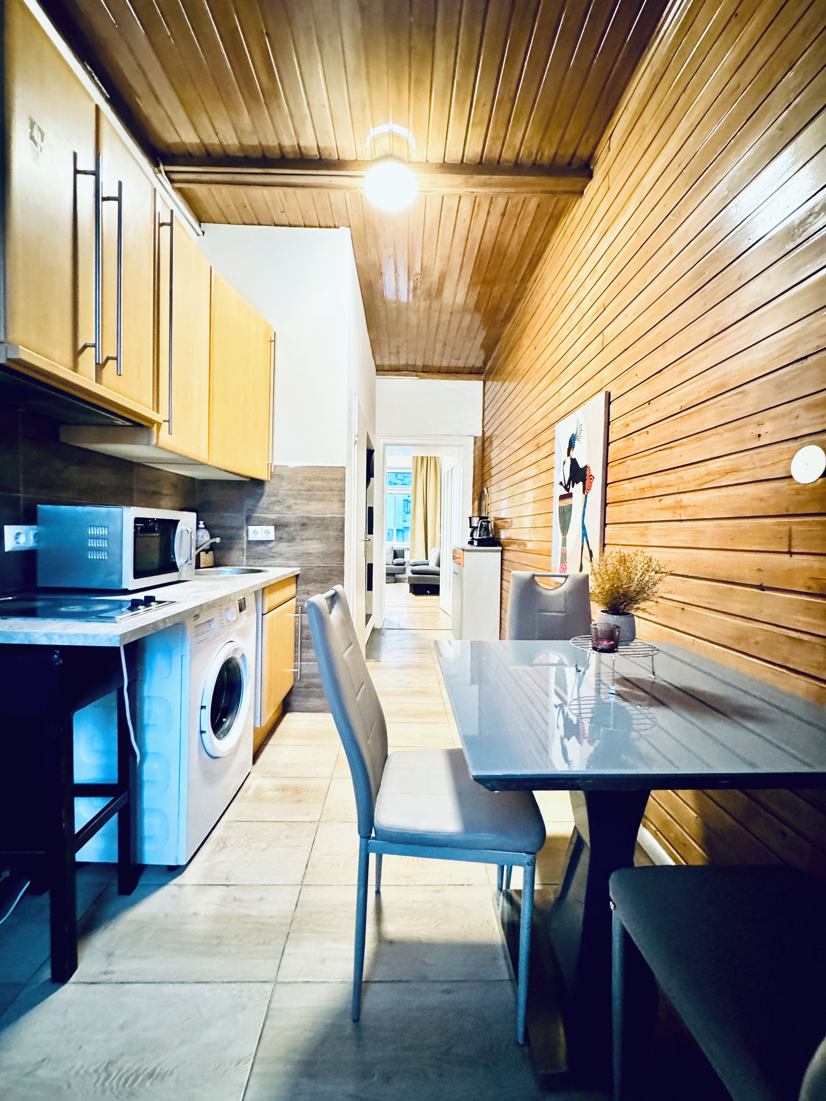 A compact kitchen with wooden cabinets, a microwave, a washing machine, and a dining area with a table and chairs.