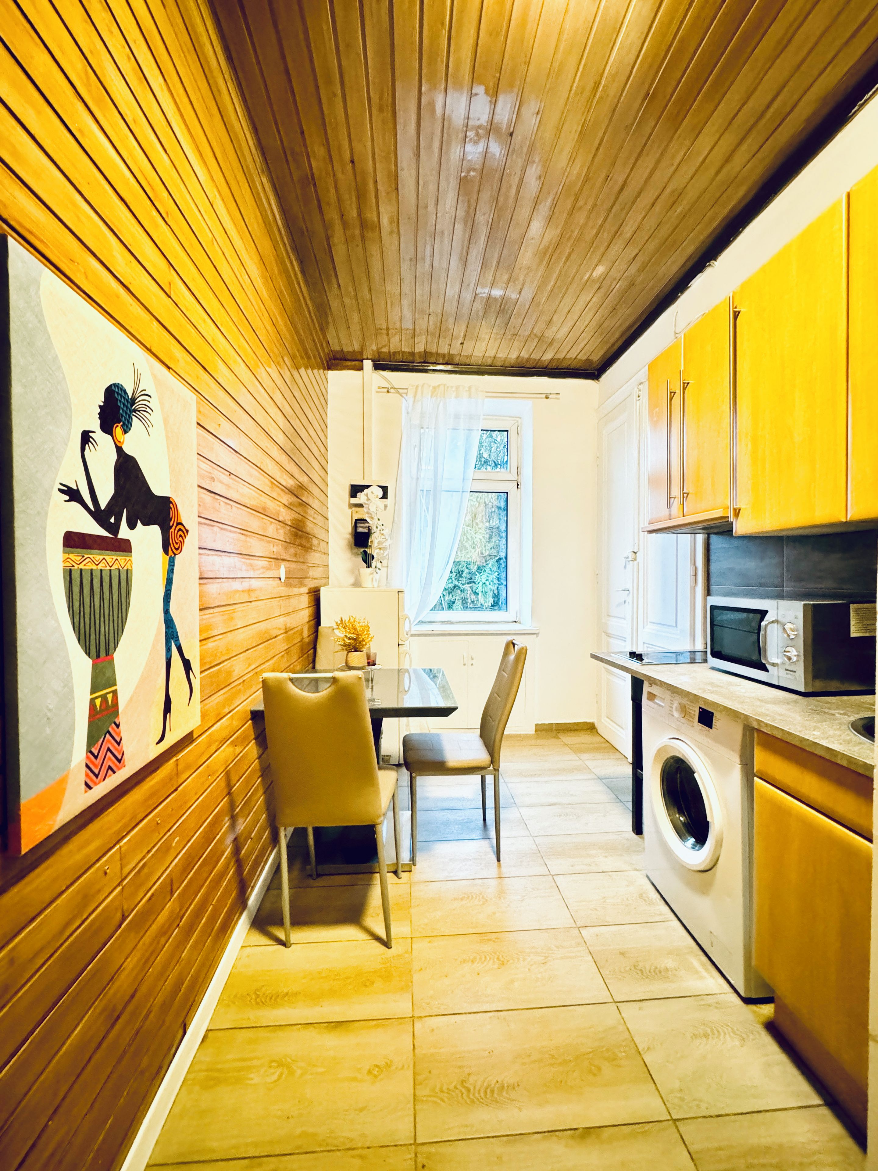 A compact kitchen with wooden walls, a dining area with chairs, a washing machine, and a microwave.