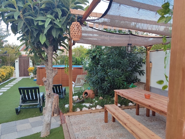 A patio area with a wooden table, a bench, a hanging light fixture, and a pergola with a mesh roof.