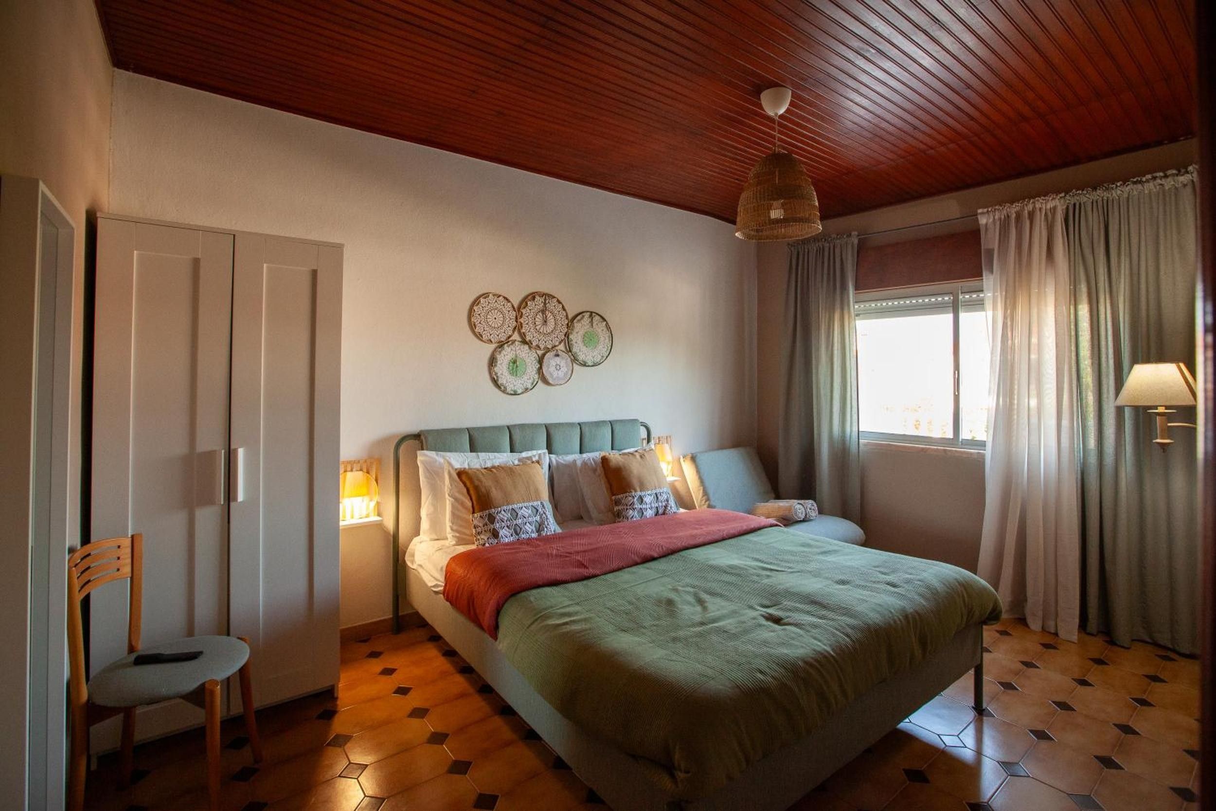 Een slaapkamer met een bed, een stoel, een raam met gordijnen, een wandlamp en een plafondlamp.