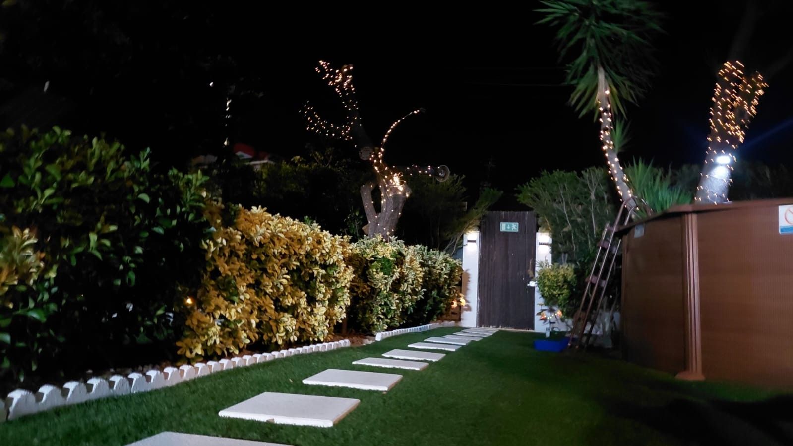 Een verlichte tuinpad 's nachts, leidend naar de ingang van een gebouw, omringd door keurig onderhouden hagen en versierd met lichtslingers.