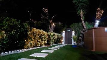 Een verlichte tuinpad 's nachts, leidend naar de ingang van een gebouw, omringd door keurig onderhouden hagen en versierd met lichtslingers.