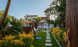 A pathway leading to a building with a tiled roof, surrounded by lush greenery and yellow flowers.