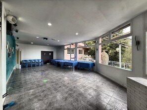 Una habitación amplia con suelo de baldosas, una mesa larga con manteles azules y una ventana con vista a la vegetación.