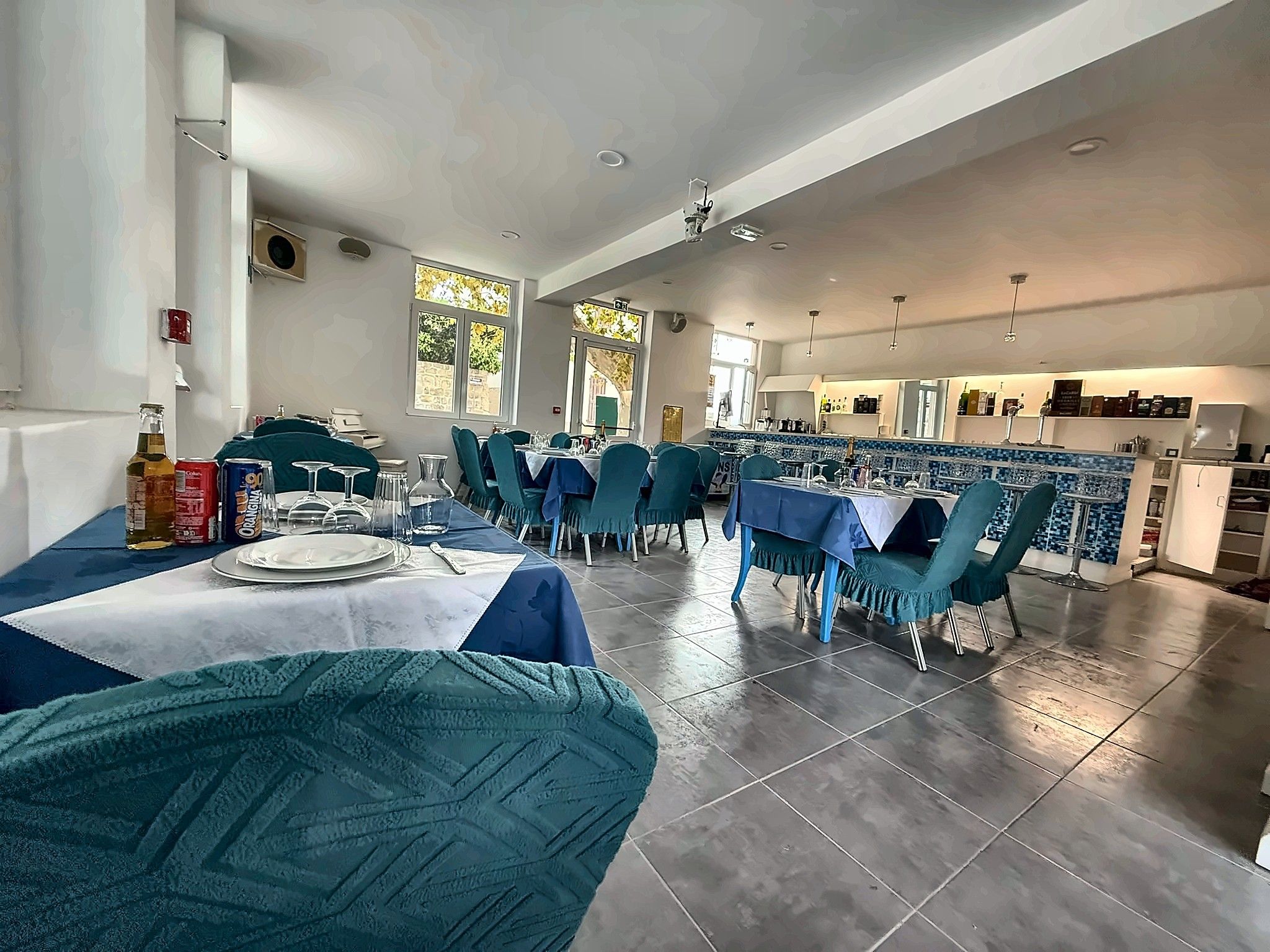 Un'ampia sala da pranzo con diversi tavoli e sedie, una cucina sullo sfondo e ampie finestre che lasciano entrare luce naturale.