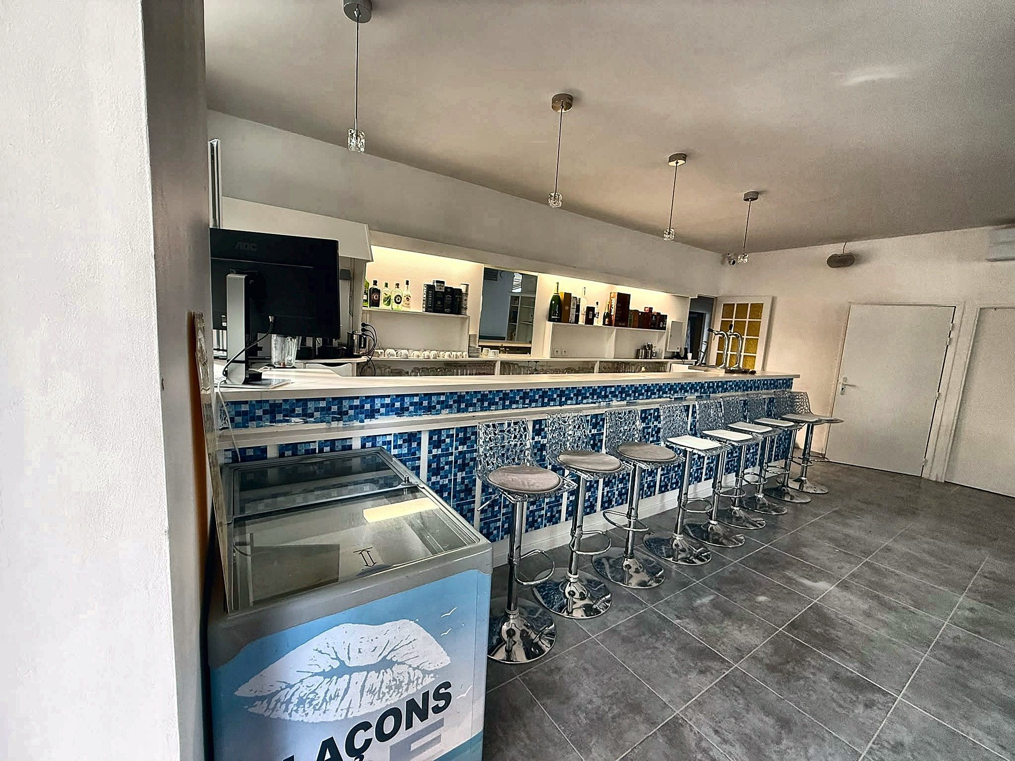 Angolo bar con schienale azzurro in piastrelle, sgabelli da bar in acciaio inox e un bancone con televisore e diverse bottiglie.