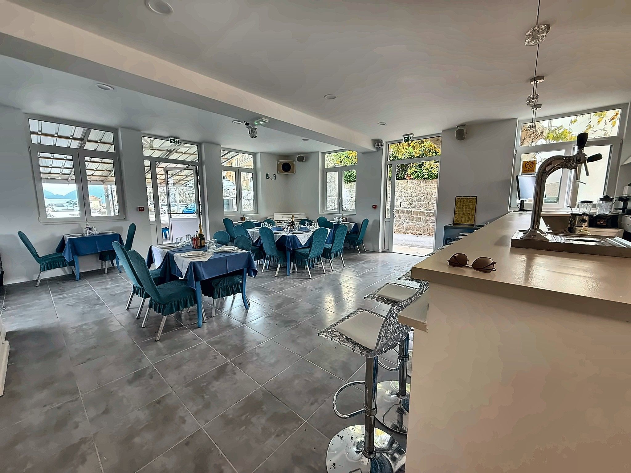 A spacious dining area with multiple tables and chairs, a bar counter with a tap, and large windows allowing natural light.