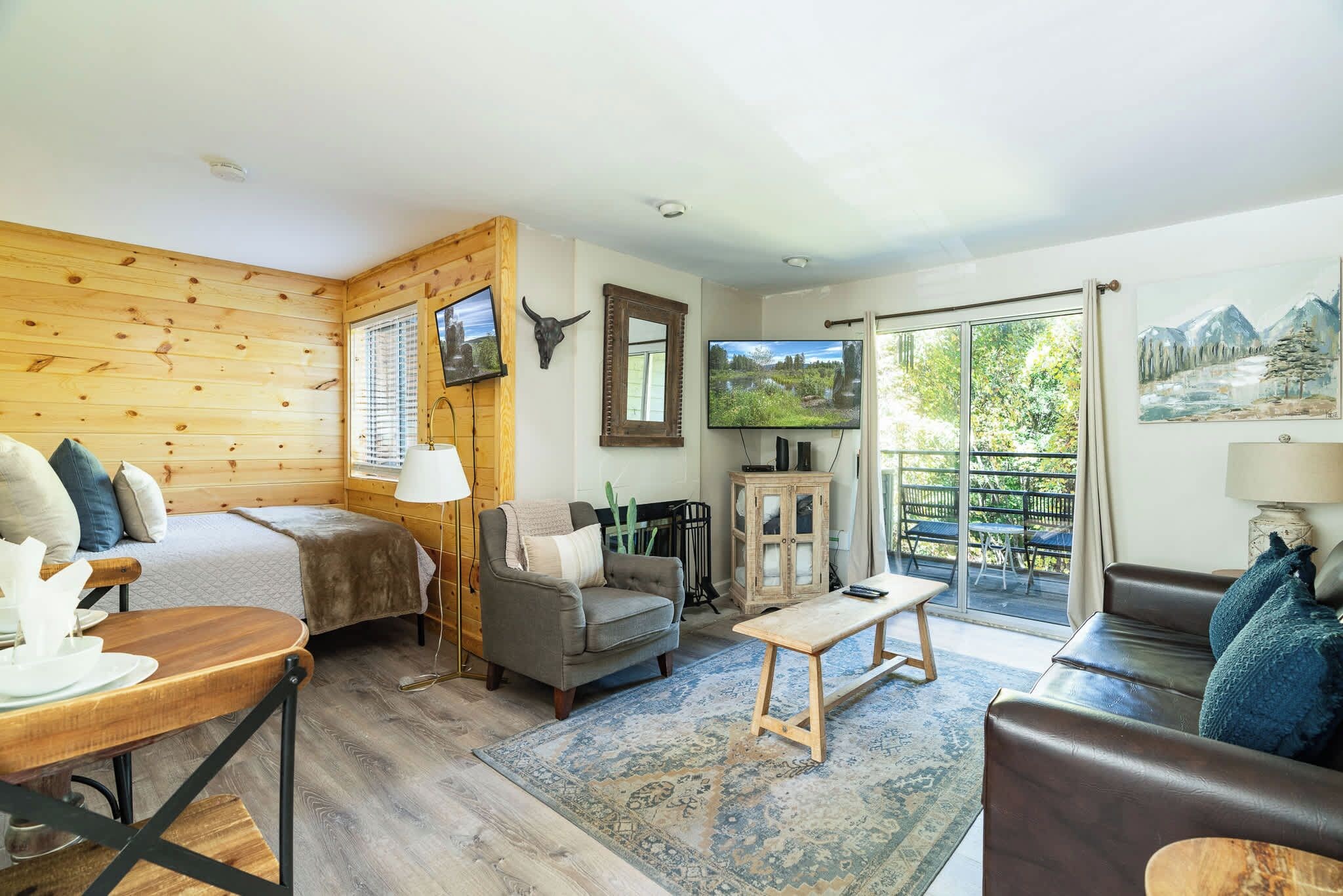 A cozy room with a bed, a sofa, a coffee table, and a TV. There is a window with blinds and a painting on the wall.
