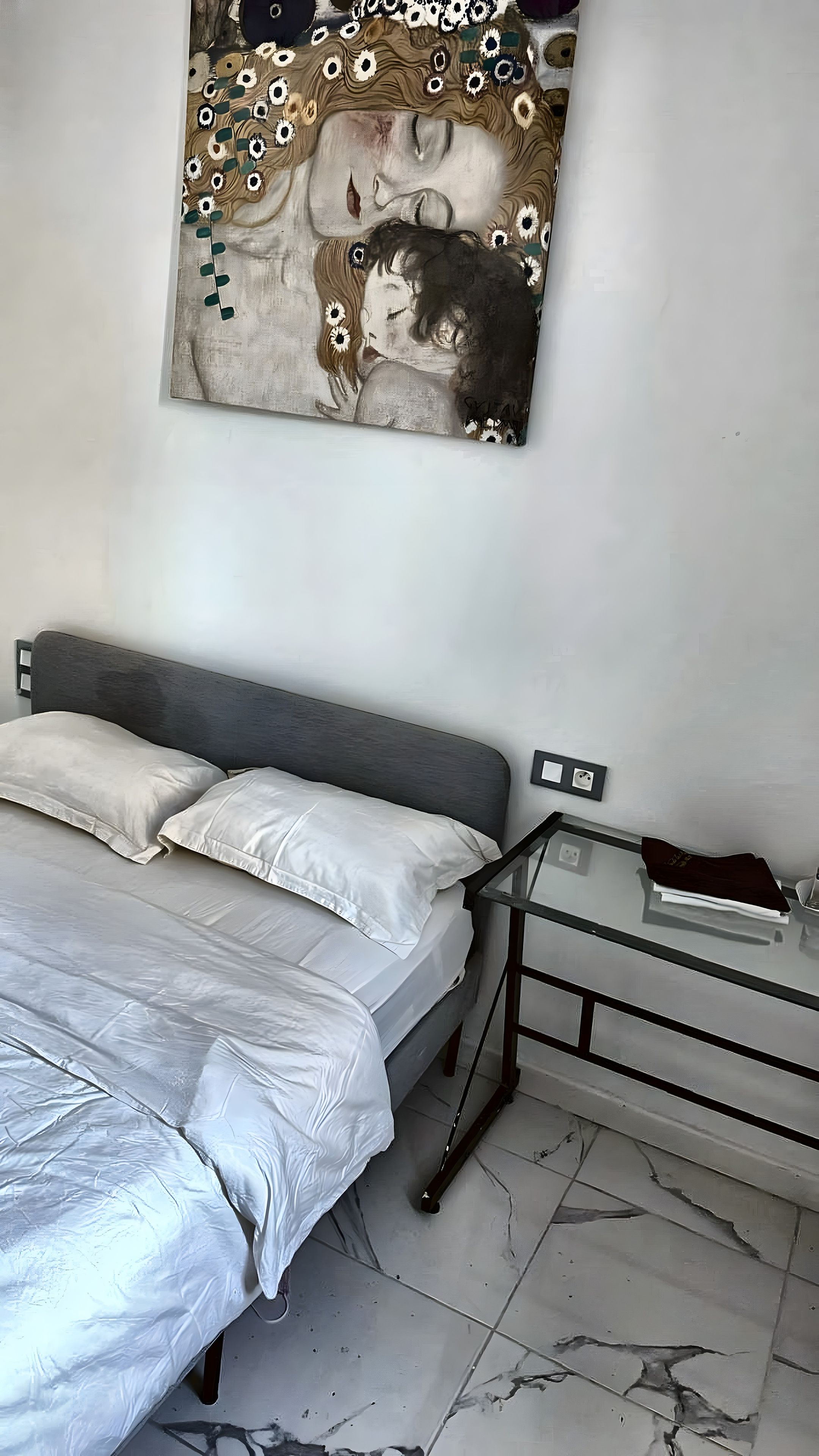 Una camera da letto con un letto, cuscini, un comodino, un quadro appeso al muro e un pavimento piastrellato.