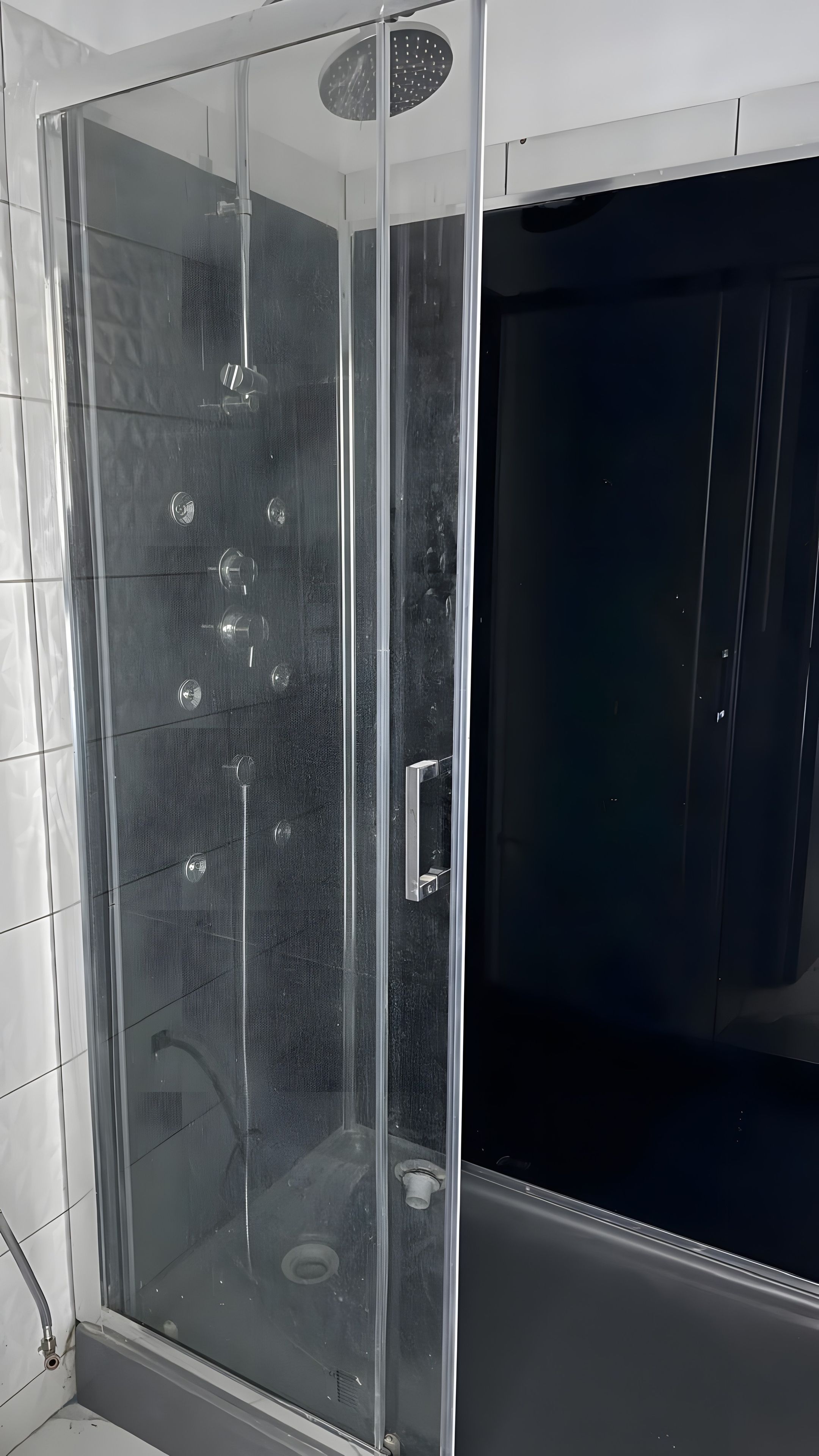 Una cabina doccia in vetro con pannello di controllo, porta scura e pareti piastrellate.
