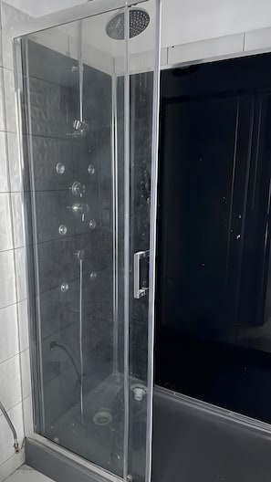 Ducha con paredes de azulejos, cabina de vidrio y panel de control, junto a una puerta oscura.