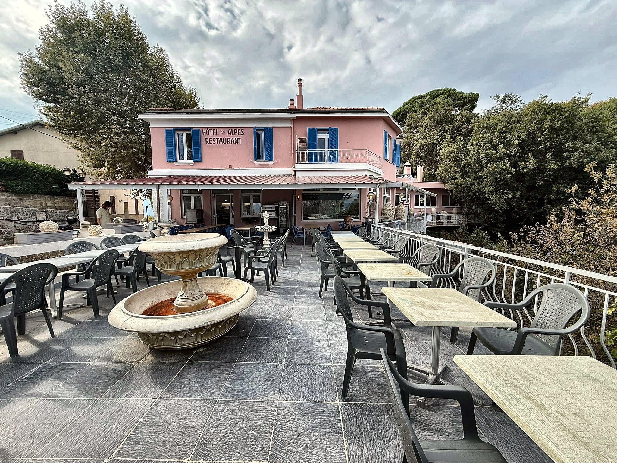 An outdoor seating area with tables and chairs, a fountain, and a building labeled 'HOTEL del ALPI RESTAURANT' in the background.