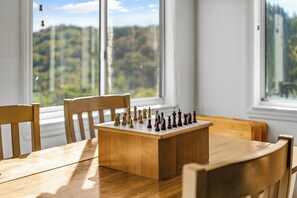 Un échiquier posé sur une table en bois, entouré de chaises, près d’une fenêtre donnant sur des arbres.