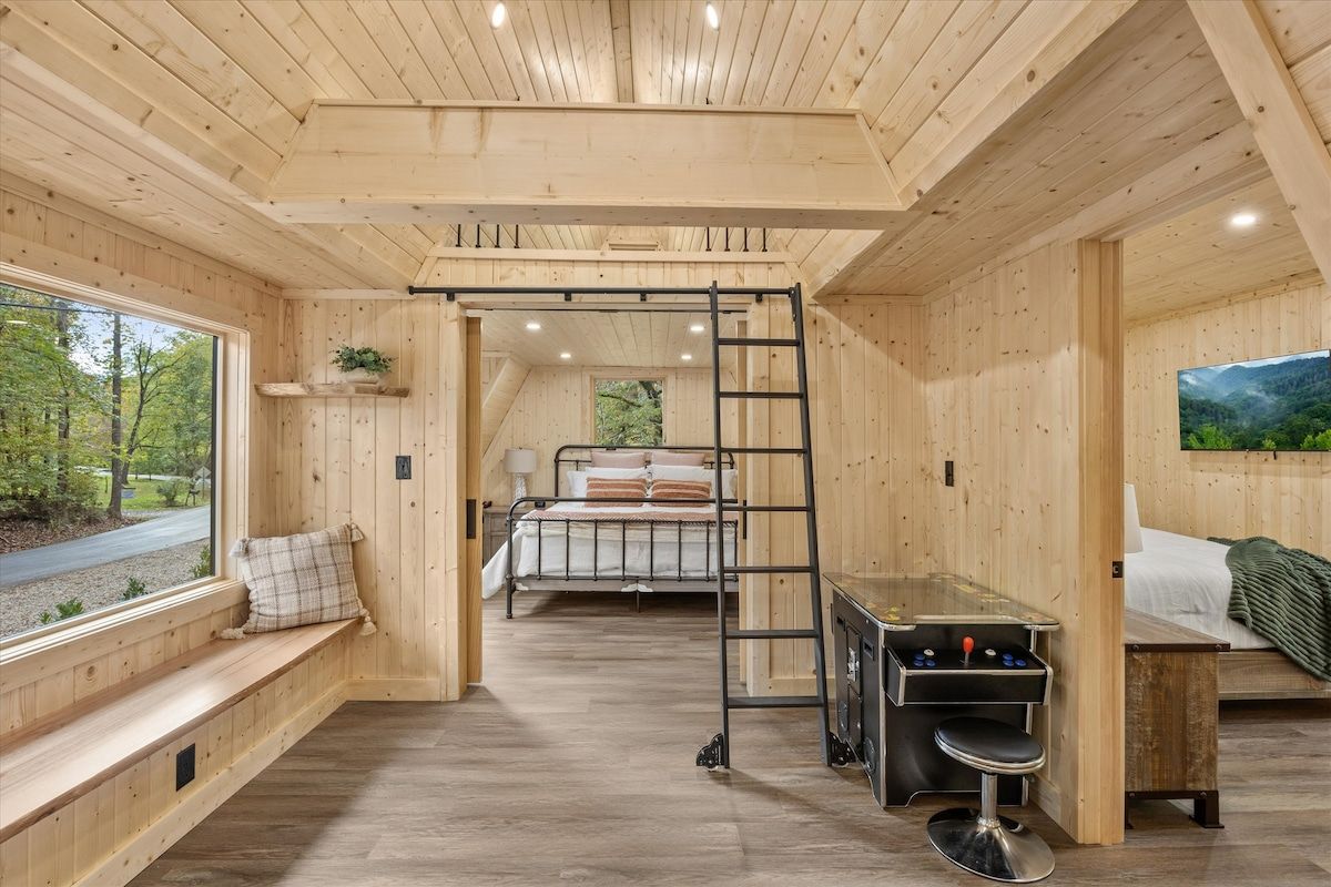 A wooden cabin interior with a bed, bench seating, a ladder, and a small table with a stool.