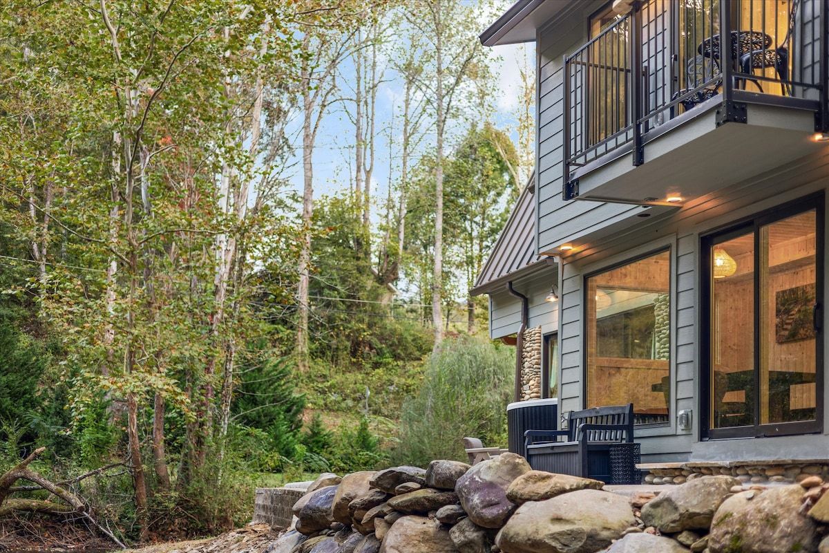 A two-story house with a stone wall in front, a balcony with outdoor furniture, and a forested hillside in the background.