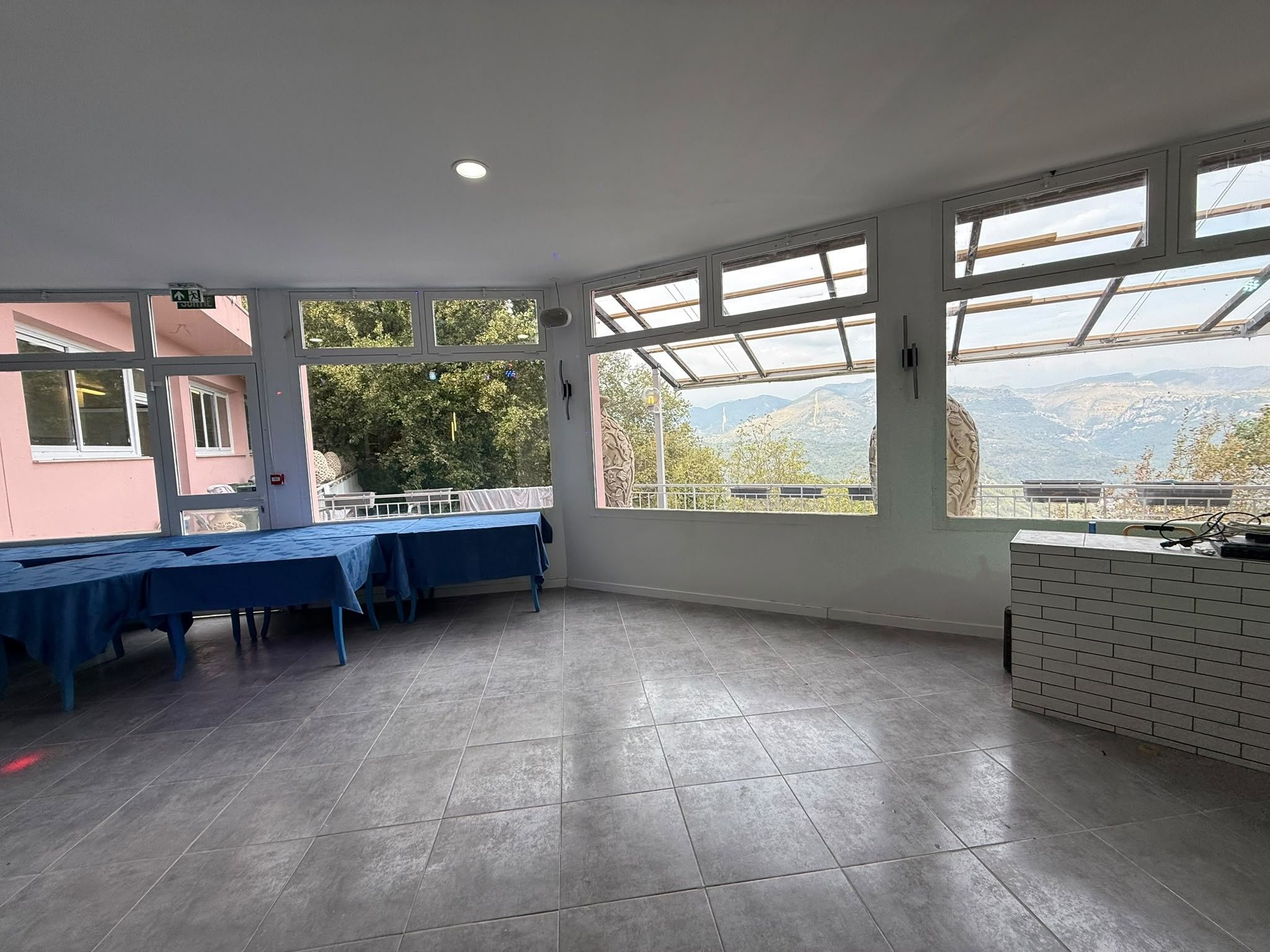 A spacious room with large windows, a tiled floor, and a table set up for dining.