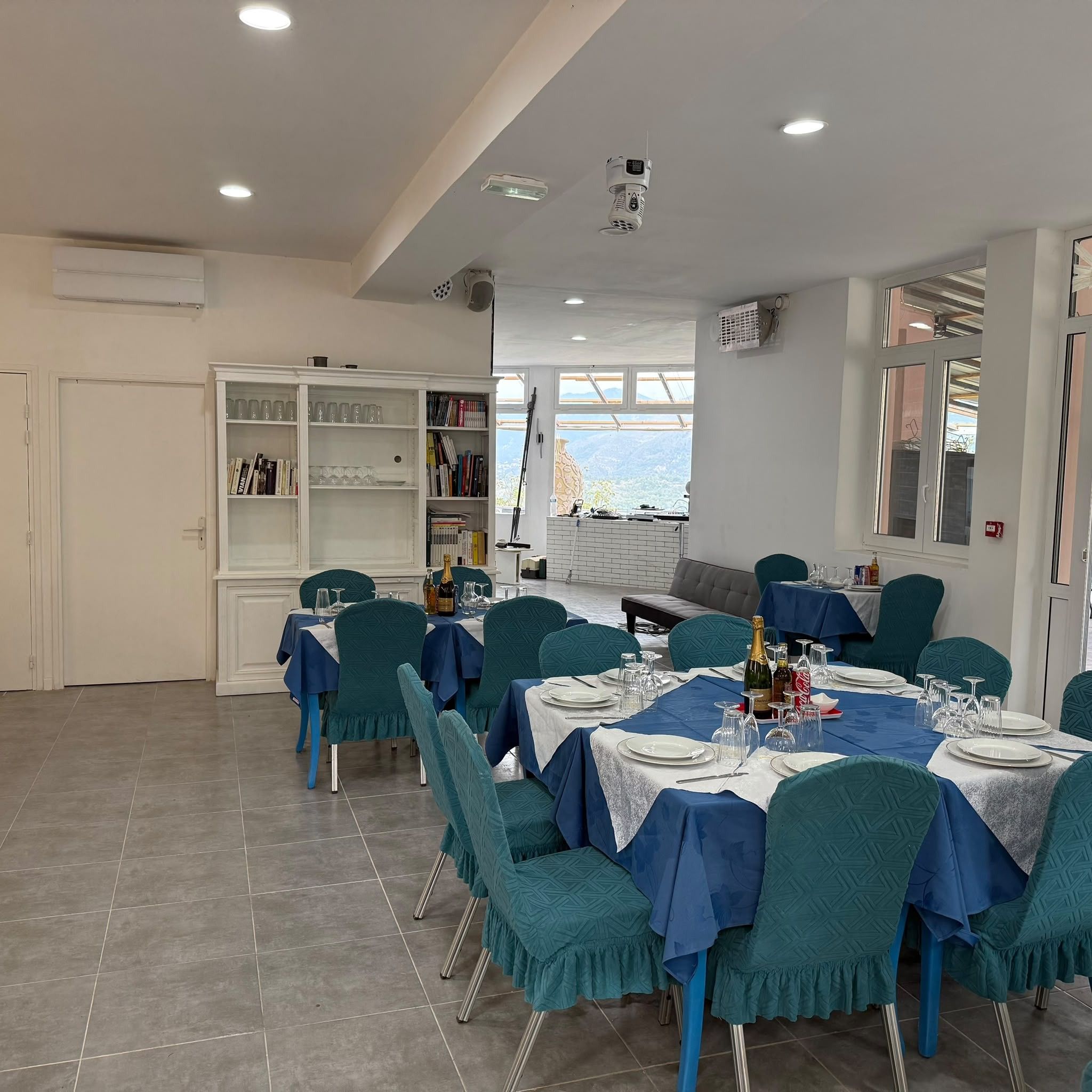 Zona pranzo con tavoli apparecchiati per quattro, sedie color petrolio e un armadietto bianco con bicchieri.