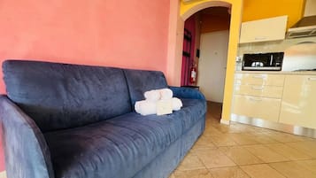Un sofá azul oscuro con una pila de toallas blancas, una zona de cocina con microondas y estufa, y una pared rosa.