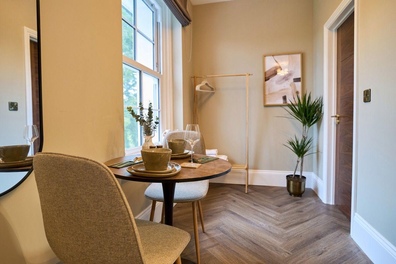 A dining area with a round table, chairs, and a vase with a plant. There is a window with blinds, a door, and a wall-mounted light fixture.