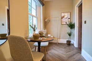 A dining area with a round table, chairs, and a vase with a plant. There is a window with blinds, a door, and a wall-mounted light fixture. - *NEW* Luxurious West Sussex Suite, Arundel Castle Views (Warningcamp)