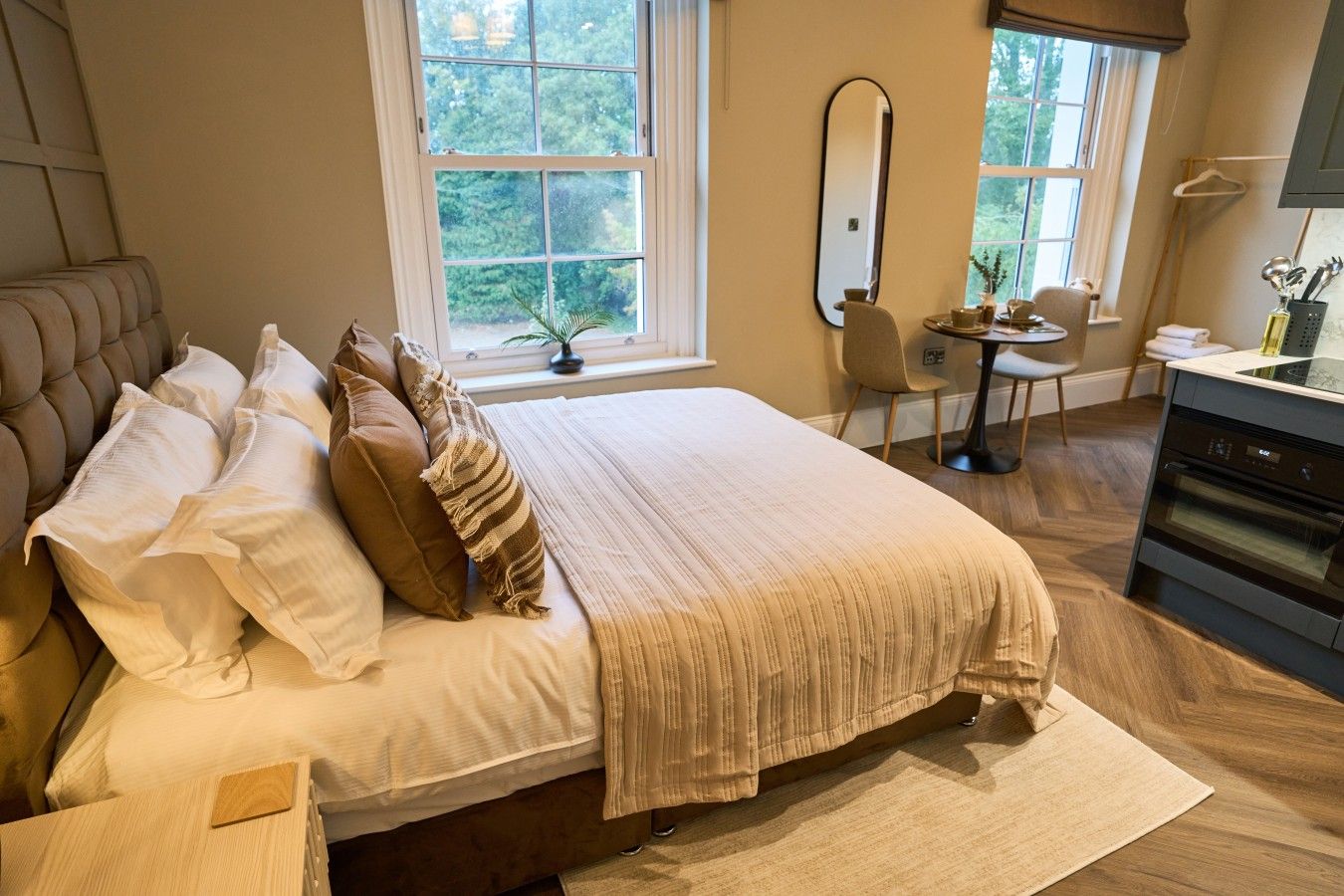 A bedroom with a large bed, a dining table, a chair, a mirror, and a kitchen area.