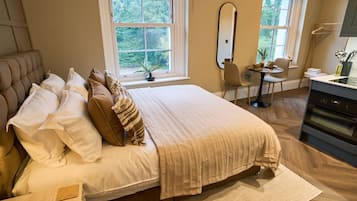 A bedroom with a large bed, a dining table, a chair, a mirror, and a kitchen area.