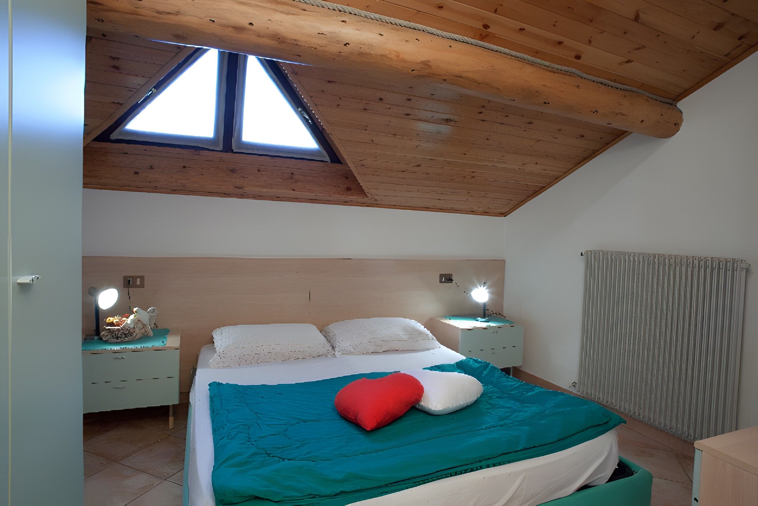A bedroom with a sloped ceiling, a skylight, a bed with a teal and white bedspread, two bedside lamps, and a nightstand with a lamp.