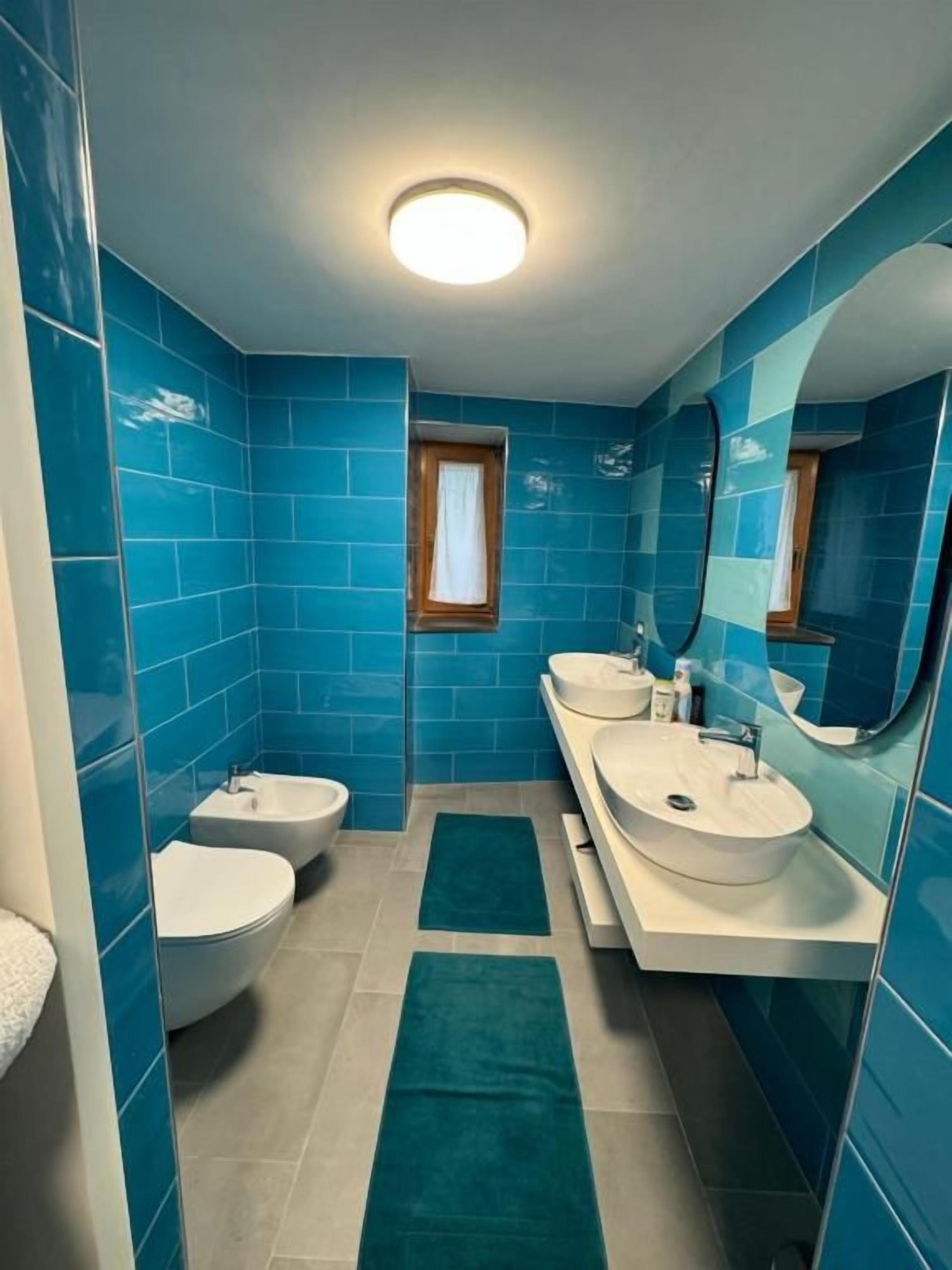 A bathroom with blue tiles, two sinks, a bidet, and a mirror.