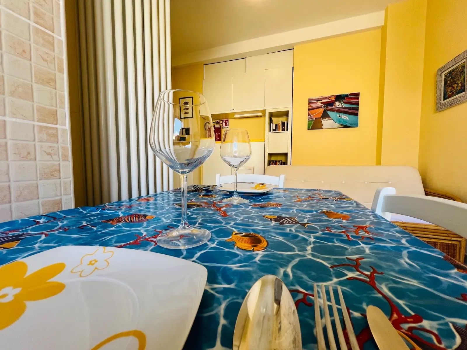 A dining area with a table set for two, featuring a blue tablecloth with fish patterns, white plates with yellow floral designs, and clear wine glasses.