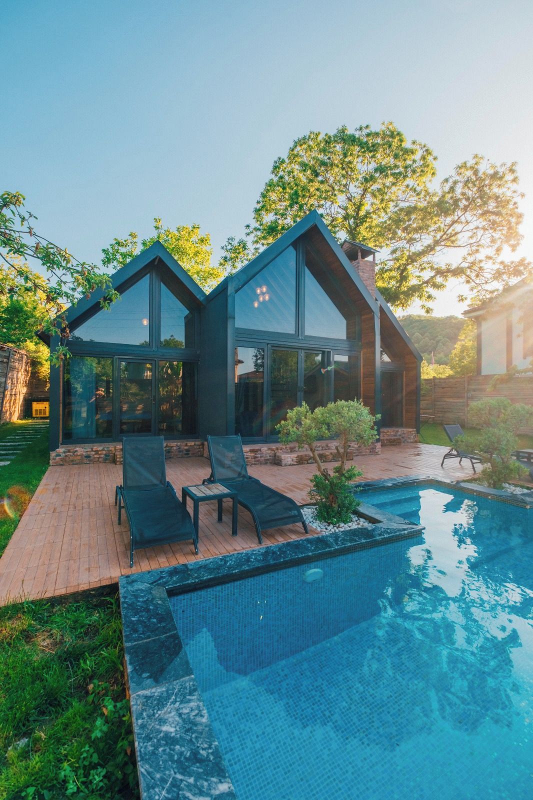 Una casa moderna con piscina, dos sillones de descanso y un pequeño jardín.