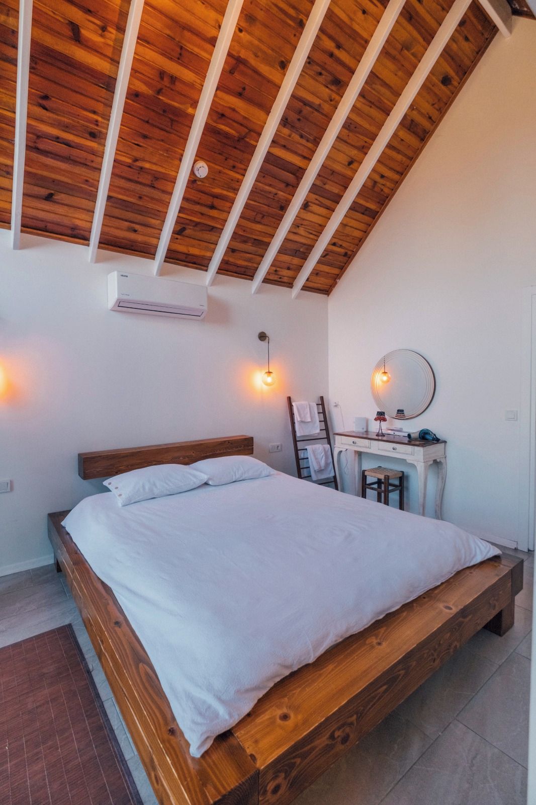 A bedroom with a wooden ceiling, a bed, a desk with a chair, and wall-mounted lighting.