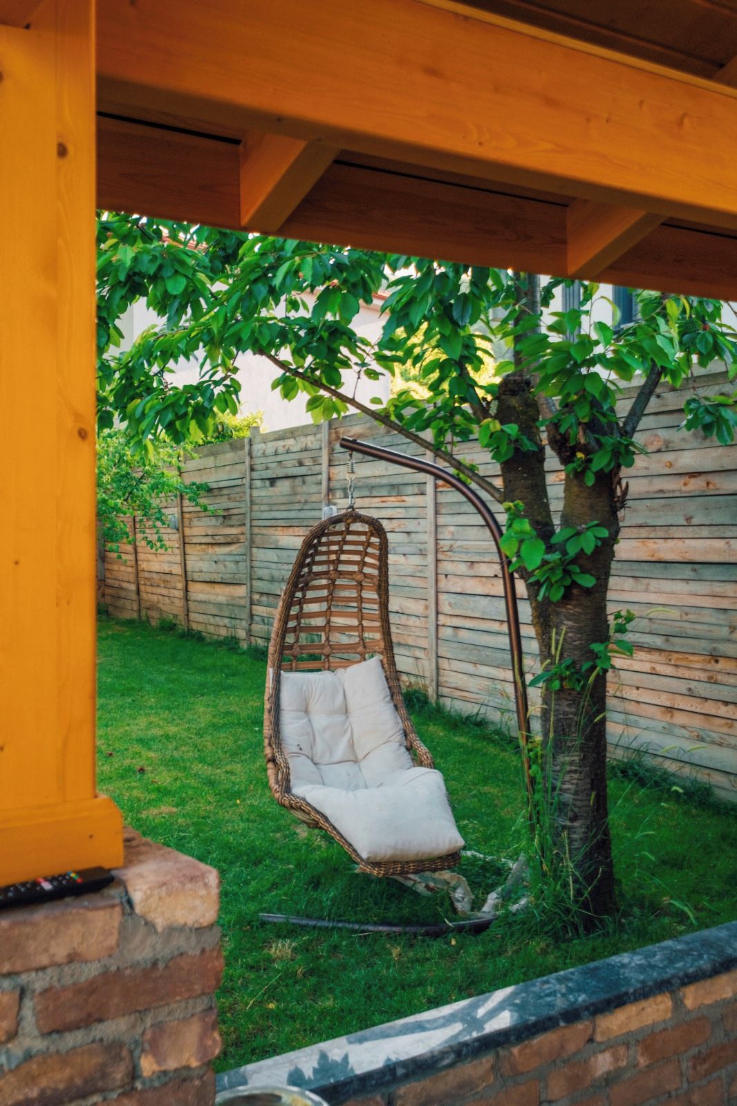 A wicker swing chair under a wooden pergola in a garden with a brick wall and a wooden fence in the background.